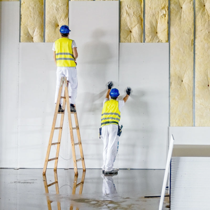 K-und-M_Trockenbau_Wärmedämmung_Isolation Zwei Trockenbau-Handwerker montieren Wärmedämmung an Innenwänden – einer arbeitet auf der Leiter, der andere hält Platten fest – fachgerechte Wärmedämmung und energetische Sanierung.